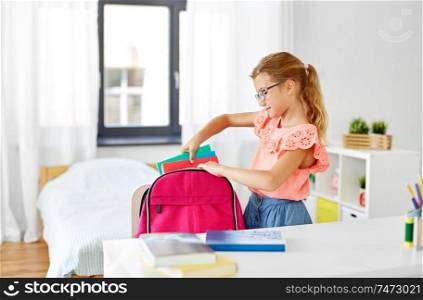 children, education and learning concept - happy student girl in glasses packing school bag at home. student girl in glasses packing school bag at home