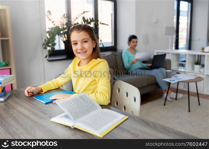 children, education and learning concept - student girl with book writing to notebook and mother working at home. student girl with book writing to notebook at home