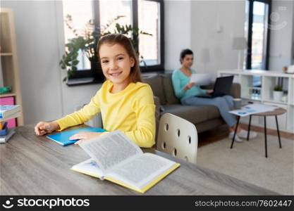 children, education and learning concept - student girl with book writing to notebook and mother working at home. student girl with book writing to notebook at home
