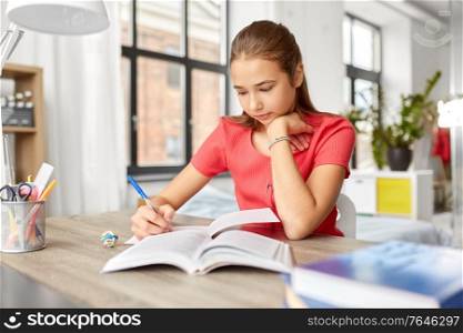 children, education and learning concept - student teenage girl with book writing to notebook at home. student girl with book writing to notebook at home