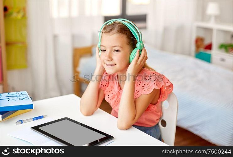 children, education and technology concept - happy student girl in headphones with tablet computer at home desk. girl in headphones with tablet computer at home