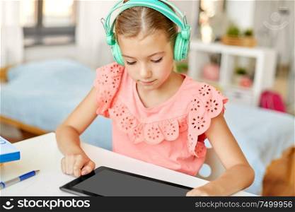children, education and technology concept - student girl in headphones with tablet computer at home desk. girl in headphones with tablet computer at home