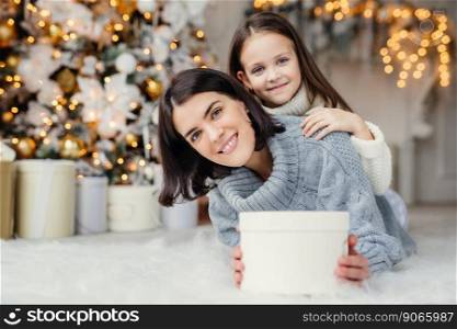 Children, family and celebration concept. Adorable female in knitted sweater holds white present box and small kid stands behind her back, have good mood before New Year s Eve. Waiting miracle