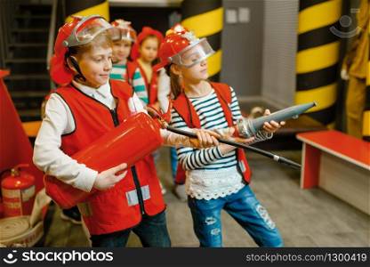 Children in helmet and uniform with hose and extinguisher in hands playing fireman, playroom indoor. Kids lerning firefighter profession. Child lifeguard, little heroes in equipment on playground. Children with hose and extinguisher, little heroes