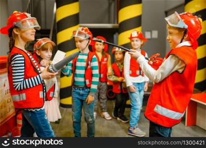 Children in helmet and uniform with hose and extinguisher in hands playing fireman, playroom indoor. Kids lerning firefighter profession. Child lifeguard, little heroes in equipment on playground