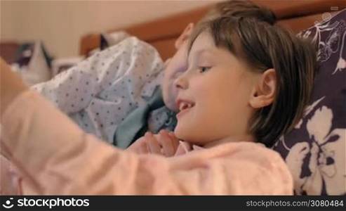 Children lying in bed and playing game on tablet PC. Lovely boy and girl kissing each other by turn