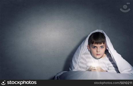 Children nightmare. Little cute boy sitting in bed under blanket with flashlight