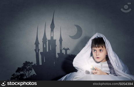 Children nightmare. Little cute boy sitting in bed under blanket with flashlight