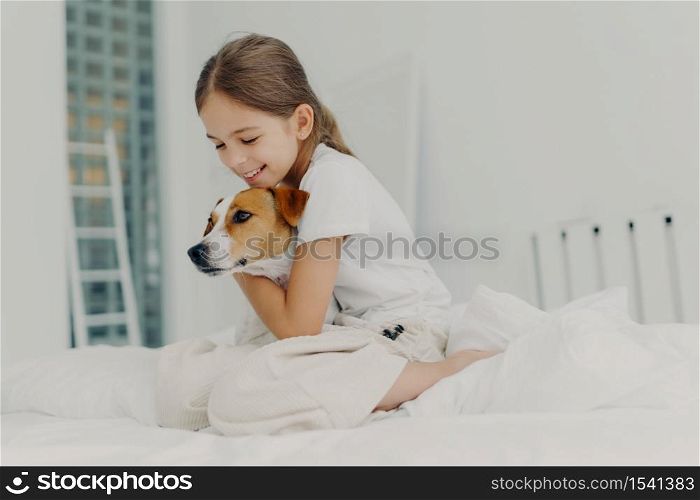 Children, rest and domestic atmosphere. Happy little female child wears nightclothes, cuddle small dog, expresses love and care to favourite pet, pose on comfortable bed with white bedclothes