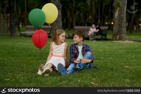 Children&rsquo;s romantic date in summer park, friendship, first love. Boy and girl with air balloons sitting back to back on a grass. Kids having fun outdoors, happy childhood. Boy and girl with balloons sitting back to back
