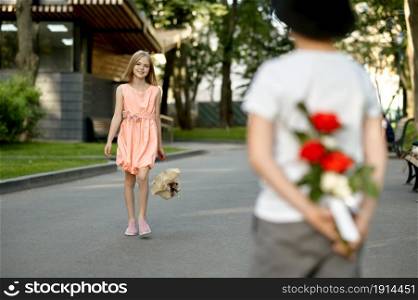 Children&rsquo;s romantic date in summer park, friendship, first love. Boy hides flowers from a girl. Kids having fun outdoors, happy childhood. Children&rsquo;s date, boy hides flowers from a girl