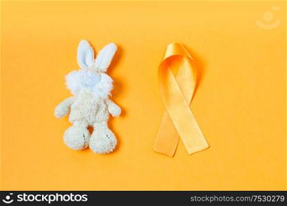 Children&rsquo;s toy rabbit with a Childhood Cancer Awareness yellow ribbon on yellow background, top view. Childhood Cancer Awareness concept
