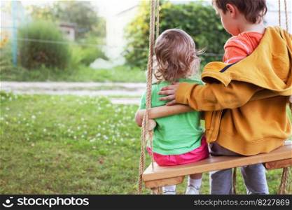 children sitting on a swing in the garden. older brother hugging little sister