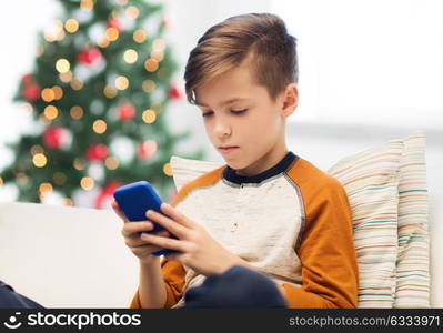 children, technology, addiction and people concept - boy with smartphone texting message or playing game at home over christmas tree background. boy with smartphone at home at christmas