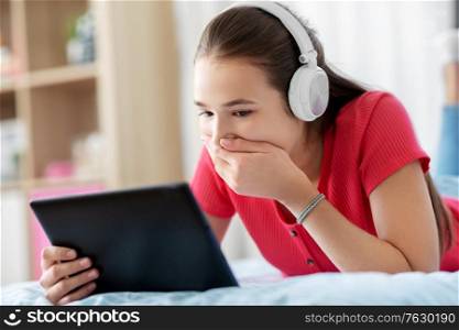 children, technology and communication concept - embarrassed teenage girl in headphones listening to music on tablet computer at home. girl in headphones listening to music on tablet pc