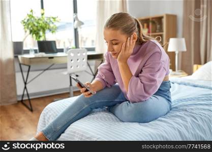children, technology and communication concept - sad teenage girl with smartphone sitting on bed at home. sad girl with smartphone sitting on bed at home