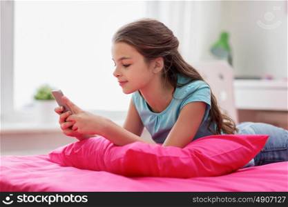children, technology and communication concept - smiling girl texting on smartphone and lying in bed at home. smiling girl texting on smartphone at home