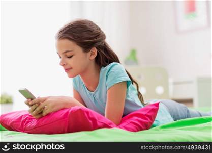 children, technology and communication concept - smiling girl texting on smartphone and lying in bed at home. smiling girl texting on smartphone at home