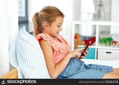 children, technology and communication concept - smiling girl using smartphone sitting on bed at home. girl using smartphone sitting on bed at home