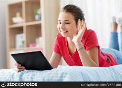 children, technology and communication concept - smiling teenage girl having vide call on tablet computer lying on bed at home. girl having vide call on tablet computer at home