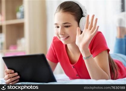 children, technology and communication concept - smiling teenage girl in headphones having vide call on tablet computer lying on bed at home. girl having vide call on tablet computer at home