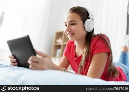 children, technology and communication concept - smiling teenage girl in headphones listening to music on tablet computer at home. girl in headphones listening to music on tablet pc
