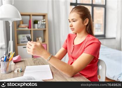 children, technology and communication concept - teenage student girl distracting from homework and texting on smartphone at home. girl with smartphone distracting from homework