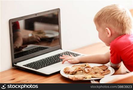 Children, technology and home concept - little boy child eating meal while using laptop pc computer at home. Bad habits