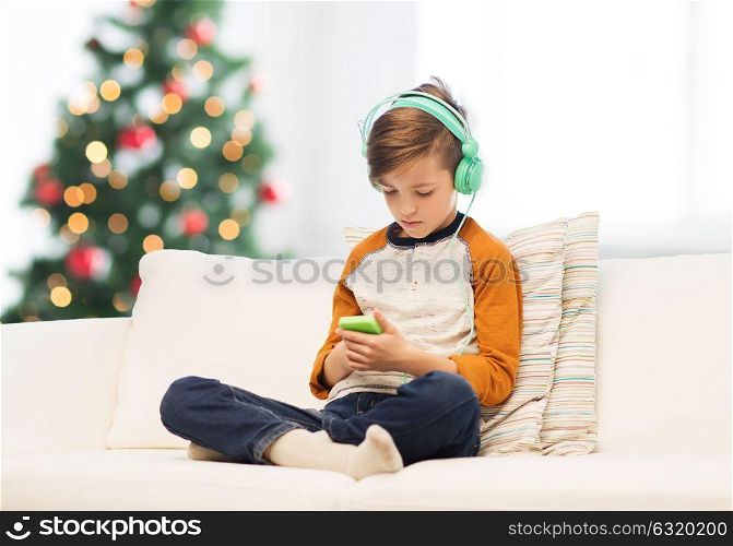 children, technology and people concept - sad boy with smartphone and headphones listening to music or playing game at home over christmas tree background. boy with smartphone and headphones at christmas