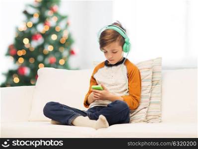 children, technology and people concept - sad boy with smartphone and headphones listening to music or playing game at home over christmas tree background. boy with smartphone and headphones at christmas