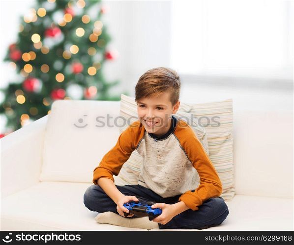children, technology and people concept - smiling boy with gamepad playing video game at home over christmas tree background. boy with gamepad playing video game at christmas
