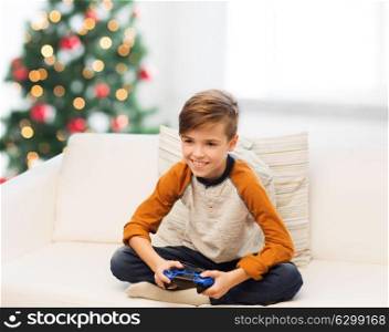 children, technology and people concept - smiling boy with gamepad playing video game at home over christmas tree background. boy with gamepad playing video game at christmas
