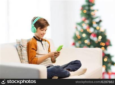 children, technology and people concept - smiling boy with smartphone and headphones listening to music at home over christmas tree background. boy with smartphone and headphones at christmas