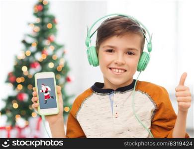 children, technology and people concept - smiling boy with smartphone and headphones listening to music and showing thumbs up at home over christmas tree background. boy with smartphone and headphones at christmas