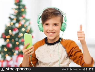 children, technology and people concept - smiling boy with smartphone and headphones listening to music at home over christmas tree background showing thumbs up. boy with smartphone and headphones at christmas