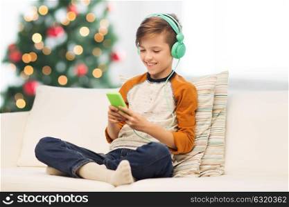 children, technology and people concept - smiling boy with smartphone and headphones listening to music at home over christmas tree background. boy with smartphone and headphones at christmas