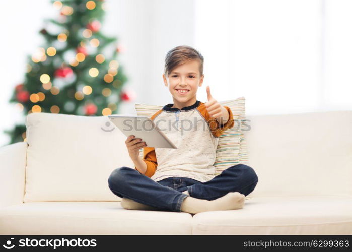 children, technology and people concept - smiling boy with tablet pc computer showing thumbs up at home over christmas tree background. boy with tablet pc showing thumbs up at christmas