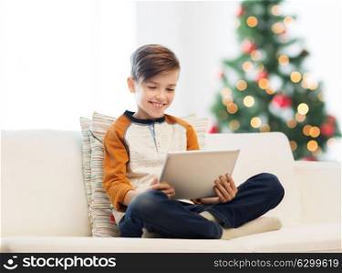 children, technology and people concept - smiling boy with tablet pc computer sitting on sofa at home over christmas tree background. smiling boy with tablet pc at home at christmas