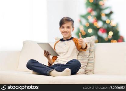 children, technology and people concept - smiling boy with tablet pc computer showing thumbs up at home over christmas tree background. boy with tablet pc showing thumbs up at christmas