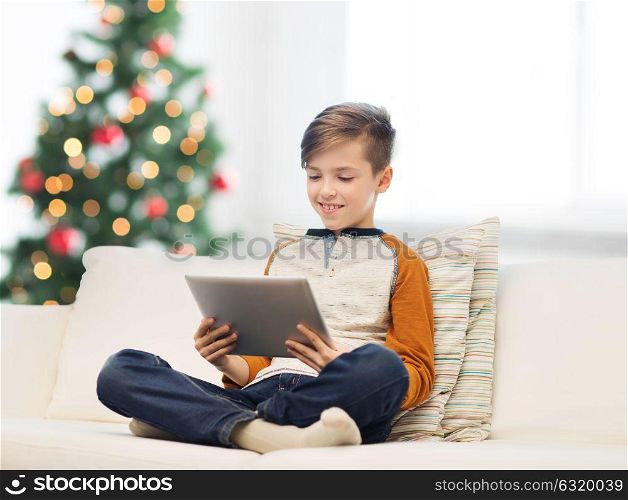 children, technology and people concept - smiling boy with tablet pc computer sitting on sofa at home over christmas tree background. smiling boy with tablet pc at home at christmas