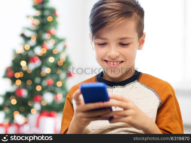 children, technology, communication and people concept - close up of smiling boy with smartphone texting message or playing game at home over christmas tree background. close up of happy boy with smartphone at christmas