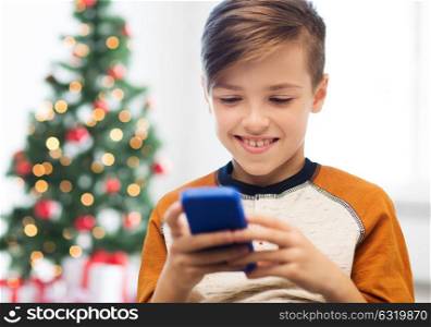 children, technology, communication and people concept - close up of smiling boy with smartphone texting message or playing game at home over christmas tree background. close up of happy boy with smartphone at christmas