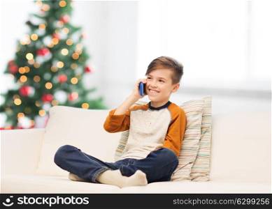 children, technology, communication and people concept - smiling boy calling on smartphone at home over christmas tree background. boy calling on smartphone at home at christmas