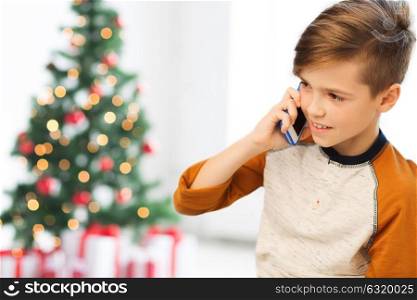 children, technology, communication and people concept - smiling boy calling on smartphone at home over christmas tree background. boy calling on smartphone at home at christmas