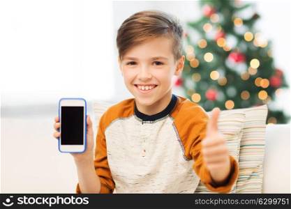 children, technology, communication and people concept - smiling boy with smartphone at home over christmas tree background showing thumbs up. boy with smartphone at christmas showing thumbs up
