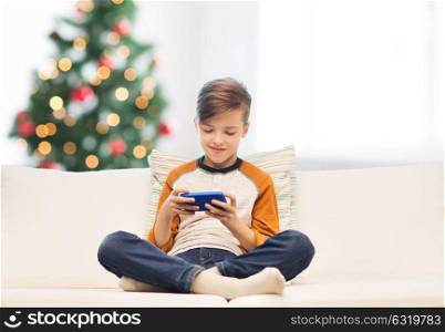 children, technology, communication and people concept - smiling boy with smartphone texting message or playing game at home over christmas tree background. boy playing on smartphone at home at christmas