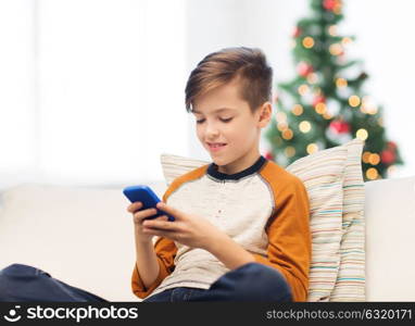 children, technology, communication and people concept - smiling boy with smartphone texting message or playing game at home over christmas tree background. boy playing on smartphone at home at christmas