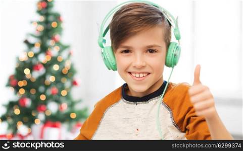 children, technology, gesture and people concept - smiling boy with headphones listening to music and showing thumbs up at home over christmas tree background. boy in headphones showing thumbs up at christmas