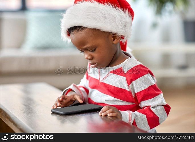 children, winter holidays and technology concept - little african american boy in santa hat with smartphone at home on christmas. african american boy with phone on christmas
