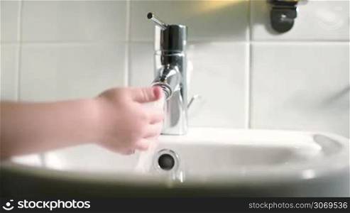Childs hand touching carefully water stream pouring from tap in the bathroom